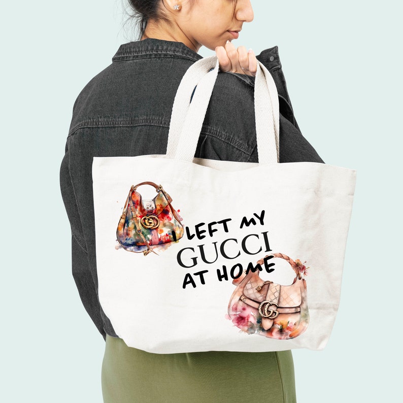 May include: White canvas tote bag with the text "LEFT MY GUCCI AT HOME" printed in black. The bag features two watercolor illustrations of Gucci handbags with floral patterns.
