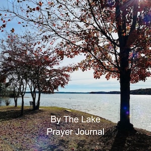 May include: A book cover with the title "By The Lake Prayer Journal" by Brittany Nidds. The cover features a scenic image of a lake with trees in the foreground and a bright blue sky.