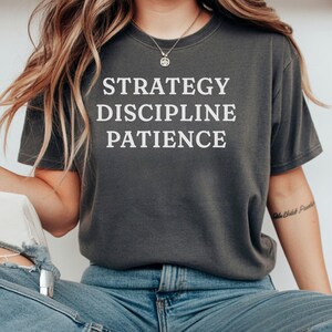 May include: Dark grey t-shirt with the words "STRATEGY DISCIPLINE PATIENCE" in white block letters. The shirt is worn with blue jeans. The overall aesthetic is casual and motivational.