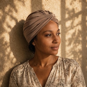 Könnte beinhalten: Ein einfarbiger, taupefarbener Turban mit einem gerafften, gedrehten Knotendetail an der Vorderseite. Der Stoff ist glatt und blickdicht und bedeckt den Kopf vollständig mit weichen, abgerundeten Falten, die ein strukturiertes Aussehen verleihen.