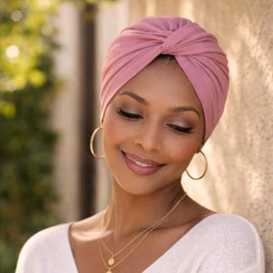 Könnte beinhalten: Ein altrosa Turban-Kopfwickel mit einem gerafften Knotendetail in der vorderen Mitte. Der Stoff ist weich und blickdicht und bedeckt den Kopf vollständig mit einer glatten, eng anliegenden Silhouette, die sicher über der Stirn sitzt.