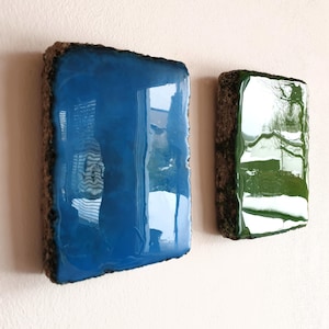 Peut inclure: Deux œuvres d'art murales abstraites. L'une est de forme rectangulaire avec une surface bleu profond brillante et une bordure marron rugueuse. L'autre est de forme similaire, mais avec une surface verte brillante et une bordure marron. Les deux sont montées sur un mur blanc.