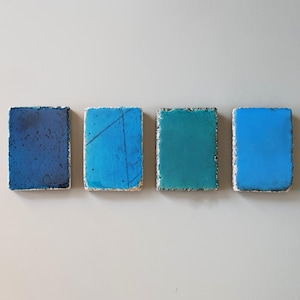 Puede incluir: Cuatro posavasos rectangulares con un borde de piedra rugoso y texturizado y una superficie superior lisa y brillante. Los colores van desde el azul marino profundo hasta el azul cielo brillante, con una fina línea diagonal oscura en uno.