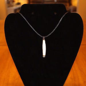 May include: A white pendant necklace with a long, thin, white pendant on a black chain. The necklace is displayed on a black velvet stand.