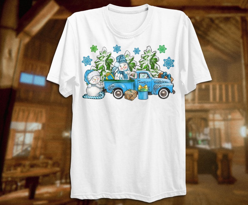 May include: A white t-shirt with a blue pickup truck carrying two snowmen and a Christmas tree. The truck is decorated with snowflakes and green pine branches.