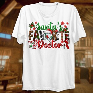 Puede incluir: Camiseta blanca con un dise&ntilde;o navide&ntilde;o que dice "El m&eacute;dico favorito de Santa". El dise&ntilde;o presenta un gorro de Pap&aacute; Noel, un estetoscopio, un botiqu&iacute;n de primeros auxilios y otros s&iacute;mbolos m&eacute;dicos.