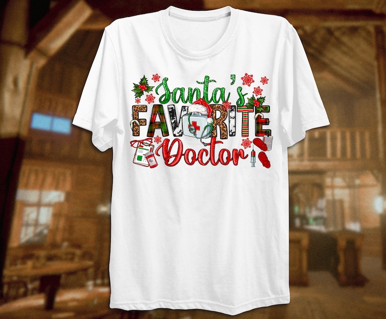 Puede incluir: Camiseta blanca con un dise&ntilde;o navide&ntilde;o que dice "El m&eacute;dico favorito de Santa". El dise&ntilde;o presenta un gorro de Pap&aacute; Noel, un estetoscopio, un botiqu&iacute;n de primeros auxilios y otros s&iacute;mbolos m&eacute;dicos.