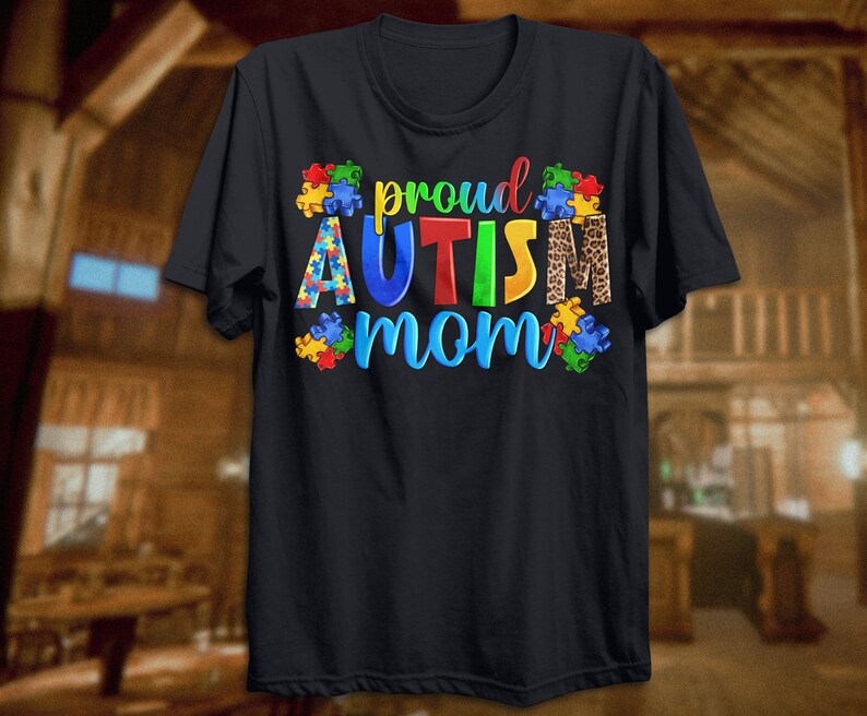 Może przedstawiać: Czarny T-shirt z napisem "proud autism mom" w kolorach tęczy z puzzlami i wzorem lamparta.