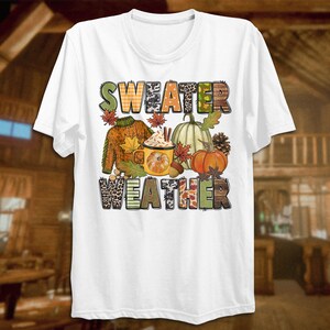 May include: White t-shirt with the text "Sweater Weather" in colorful letters. The letters are decorated with fall-themed patterns like plaid, leopard print, and leaves. The design also includes a mug of hot chocolate, pumpkins, and pinecones.
