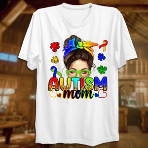 May include: White t-shirt with a colorful illustration of a woman with a messy bun, wearing glasses and a rainbow headband. The text "Autism mom" is written in rainbow letters with puzzle pieces and a ribbon.