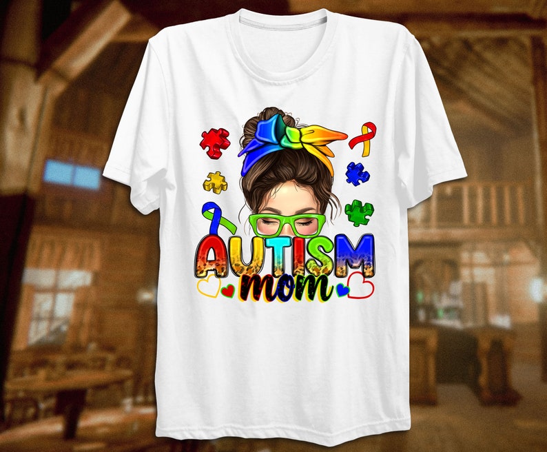 May include: White t-shirt with a colorful illustration of a woman with a messy bun, wearing glasses and a rainbow headband. The text "Autism mom" is written in rainbow letters with puzzle pieces and a ribbon.
