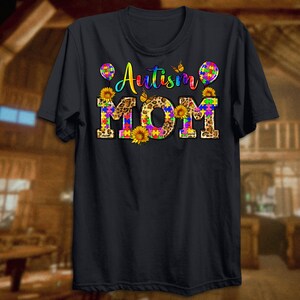 Puede incluir: Camiseta negra con el texto "Autism Mom" en piezas de rompecabezas arco&iacute;ris sobre un fondo de estampado de leopardo. El texto est&aacute; rodeado de mariposas y girasoles.