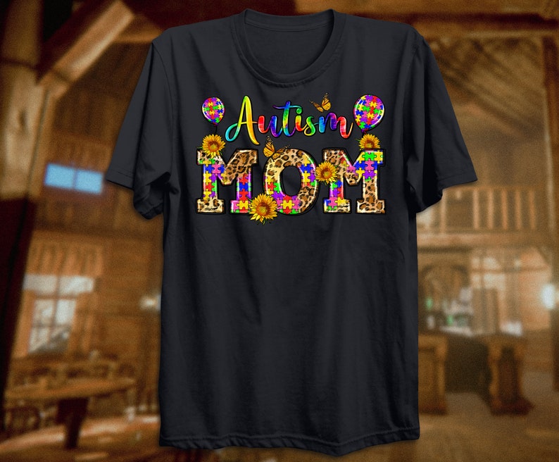 Puede incluir: Camiseta negra con el texto "Autism Mom" en piezas de rompecabezas arco&iacute;ris sobre un fondo de estampado de leopardo. El texto est&aacute; rodeado de mariposas y girasoles.