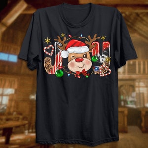 Pu&ograve; includere: Maglietta nera con un design natalizio che presenta una renna dei cartoni animati con un cappello di Babbo Natale e circondata da bastoncini di zucchero, ornamenti e luci. La parola "GIOIA" &egrave; scritta con le corna della renna e altri elementi natalizi.