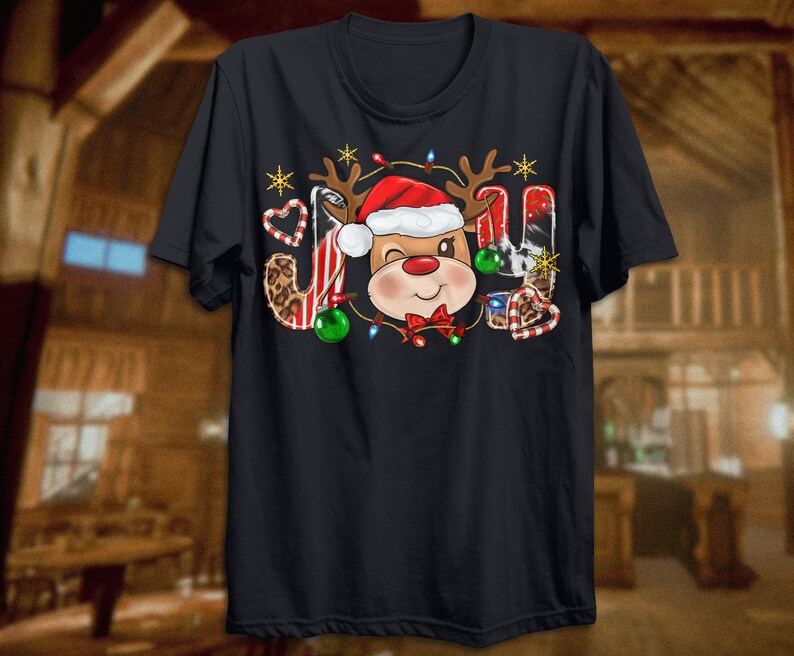 Pu&ograve; includere: Maglietta nera con un design natalizio che presenta una renna dei cartoni animati con un cappello di Babbo Natale e circondata da bastoncini di zucchero, ornamenti e luci. La parola "GIOIA" &egrave; scritta con le corna della renna e altri elementi natalizi.