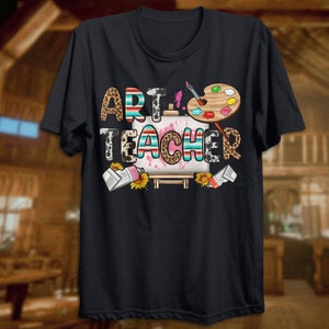 May include: Black t-shirt with the text "Art Teacher" in colorful letters with a paintbrush, paint splatters, and a paint palette on a canvas.