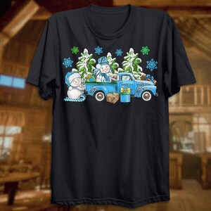 May include: A blue pickup truck with a snowman and a Christmas tree in the back. The truck is decorated with snowflakes and green pine branches. The truck is on a black t-shirt.