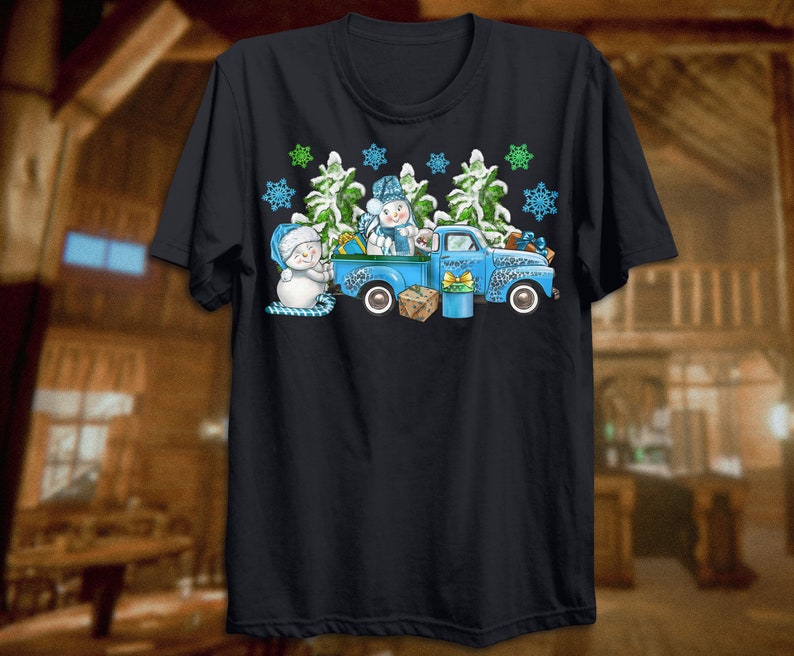 May include: A blue pickup truck with a snowman and a Christmas tree in the back. The truck is decorated with snowflakes and green pine branches. The truck is on a black t-shirt.