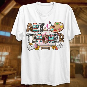 May include: White t-shirt with the text "ART TEACHER" in colorful letters with a paint palette and paintbrushes. The letters have a leopard print and striped pattern.