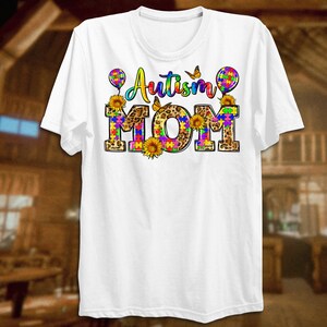 Puede incluir: Camiseta blanca con el texto "Autism Mom" en piezas de rompecabezas de colores e impresi&oacute;n de leopardo. El texto est&aacute; rodeado de mariposas, girasoles y globos.
