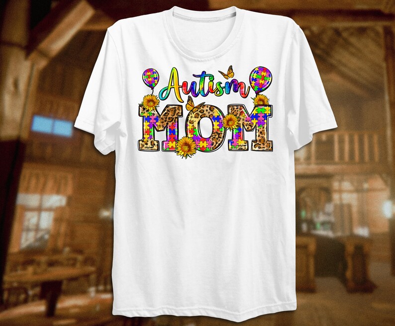 Puede incluir: Camiseta blanca con el texto "Autism Mom" en piezas de rompecabezas de colores e impresi&oacute;n de leopardo. El texto est&aacute; rodeado de mariposas, girasoles y globos.