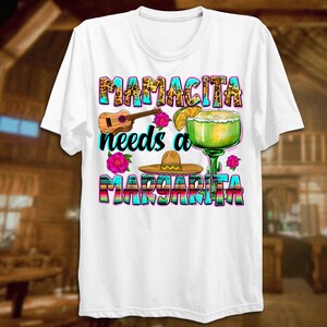 K&ouml;nnte beinhalten: Wei&szlig;es T-Shirt mit einem bunten Grafikdesign, auf dem "Mamacita needs a Margarita" steht. Die Grafik enth&auml;lt ein Margarita-Glas, einen Sombrero, eine Ukulele und Blumen.
