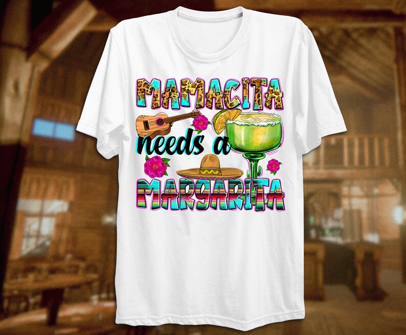 K&ouml;nnte beinhalten: Wei&szlig;es T-Shirt mit einem bunten Grafikdesign, auf dem "Mamacita needs a Margarita" steht. Die Grafik enth&auml;lt ein Margarita-Glas, einen Sombrero, eine Ukulele und Blumen.