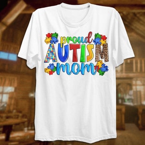 Może przedstawiać: Biała koszulka z napisem "proud autism mom" w kolorowych literach otoczonych puzzlami. Słowo "autism" jest w kolorze czerwonym, ż&oacute;łtym, niebieskim i zielonym. Elementy puzzli są niebieskie, czerwone, ż&oacute;łte i zielone. Słowo "mom" jest w kolorze niebieskim i zielonym. Za słowem "autism" znajduje się wz&oacute;r w panterkę.
