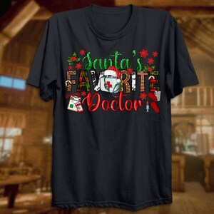 Puede incluir: Camiseta negra con un dise&ntilde;o navide&ntilde;o con el texto "El m&eacute;dico favorito de Santa" con un gorro de Pap&aacute; Noel, un botiqu&iacute;n de primeros auxilios y otros elementos navide&ntilde;os.