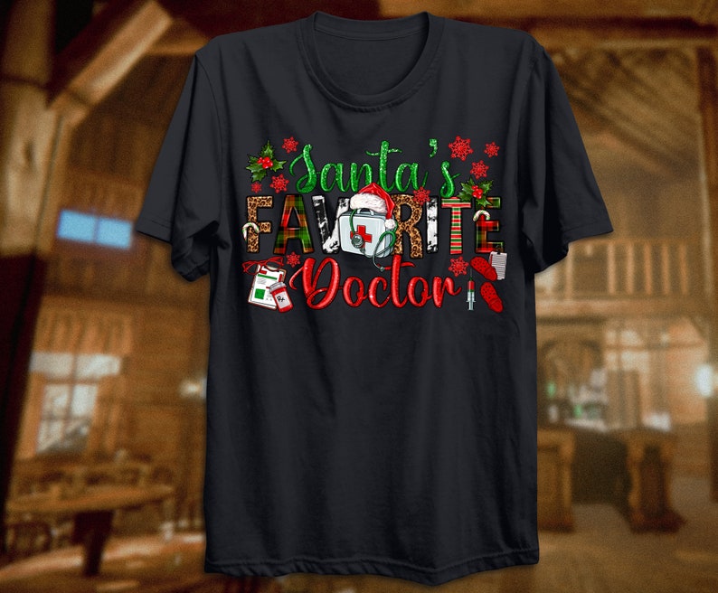 Puede incluir: Camiseta negra con un dise&ntilde;o navide&ntilde;o con el texto "El m&eacute;dico favorito de Santa" con un gorro de Pap&aacute; Noel, un botiqu&iacute;n de primeros auxilios y otros elementos navide&ntilde;os.