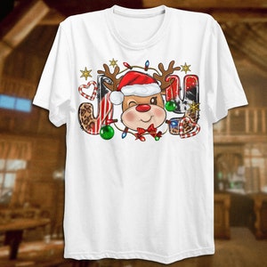 Pu&ograve; includere: Maglietta bianca con un design natalizio che presenta una renna dei cartoni animati con un cappello di Babbo Natale e circondata da bastoncini di zucchero, luci e una stampa leopardata. La parola "JOY" &egrave; scritta con la testa della renna al centro.