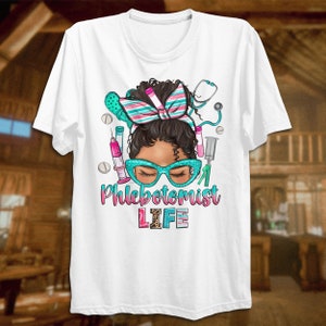 Puede incluir: Camiseta blanca con un gr&aacute;fico de una mujer negra con un mo&ntilde;o, que lleva gafas y est&aacute; rodeada de herramientas m&eacute;dicas. El texto "Phlebotomist LIFE" est&aacute; debajo del gr&aacute;fico.