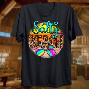 May include: Black t-shirt with a graphic of a sunset with the text "Salty Beach" in a leopard print circle. Sunglasses are in the foreground.
