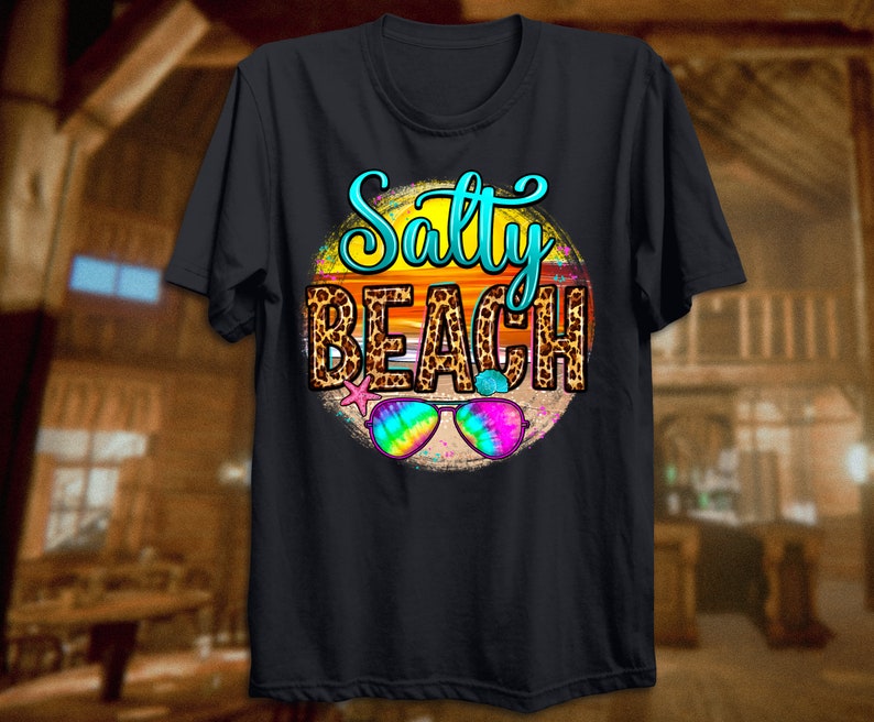 May include: Black t-shirt with a graphic of a sunset with the text "Salty Beach" in a leopard print circle. Sunglasses are in the foreground.
