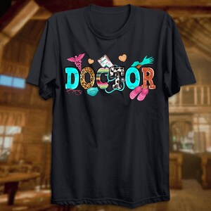 Może przedstawiać: Czarny T-shirt z napisem "DOCTOR" w kolorowych literach, ozdobiony symbolami medycznymi, stetoskopem, maską na twarz i parą r&oacute;żowych klapek.