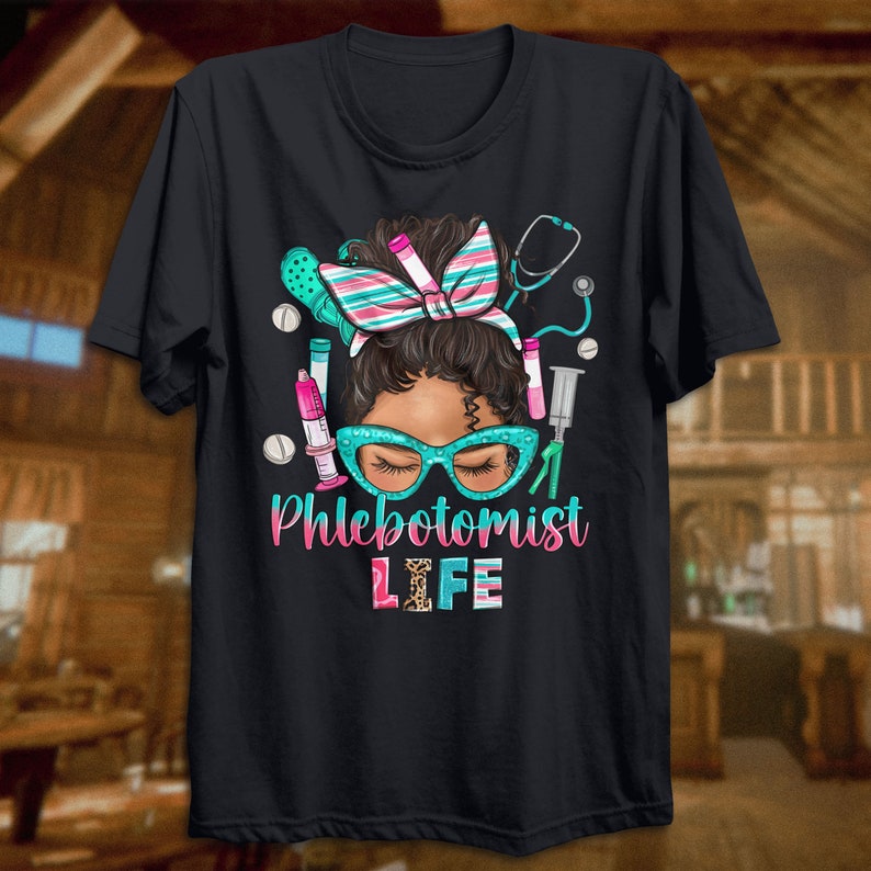 Puede incluir: Camiseta negra con una ilustraci&oacute;n de dibujos animados de una mujer con un estetoscopio, jeringas y tubos de ensayo. El texto "Phlebotomist Life" est&aacute; impreso en letras rosas y negras.