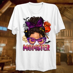 May include: White t-shirt with a graphic of a woman with a purple headband and glasses. The graphic also includes a black cat, a pumpkin, and a wand. The text "MOMSTER" is printed below the graphic.