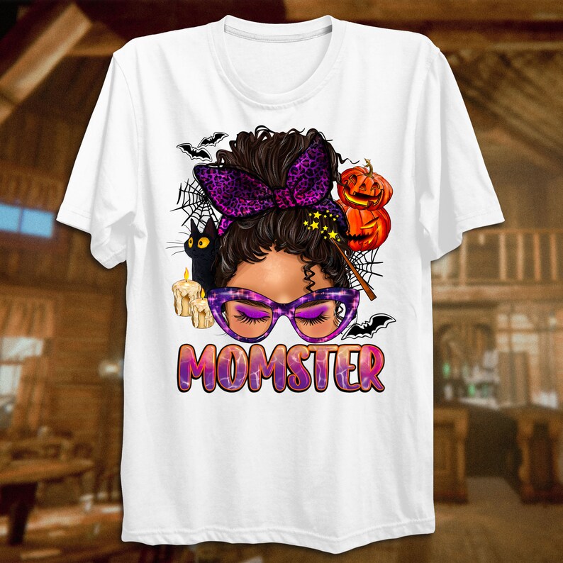 May include: White t-shirt with a graphic of a woman with a purple headband and glasses. The graphic also includes a black cat, a pumpkin, and a wand. The text "MOMSTER" is printed below the graphic.