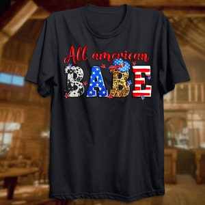 May include: A black t-shirt with the text "All American Babe" in white. The letters are decorated with red, white, and blue stars, cow print, leopard print, and a cowboy hat.