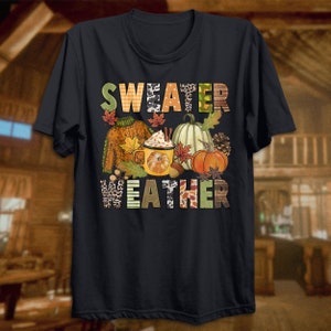 May include: Black t-shirt with a colorful graphic design that says "Sweater Weather". The graphic includes a sweater, a mug of hot chocolate, pumpkins, and fall leaves.