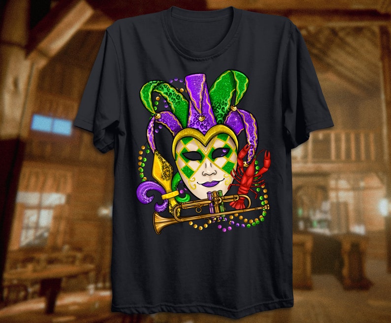 May include: A black t-shirt with a Mardi Gras theme. The design features a jester mask with green, purple, and gold accents, a trumpet, and a crawfish.