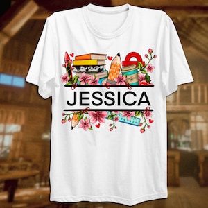 May include: White t-shirt with a colorful design of books, pencils, a ruler, a coffee cup, and flowers. The name "JESSICA" is printed in black letters on the shirt.