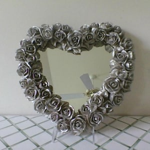 May include: A heart-shaped mirror with a silver rose frame. The mirror is supported by two small silver stands. The frame is made of many small silver roses. The mirror is on a white tiled surface.
