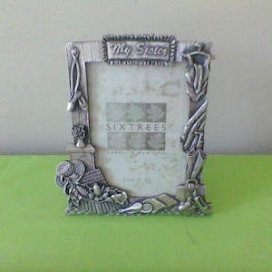 May include: A silver picture frame with a decorative design featuring a garden theme. The frame has the words "My Sister" and "Six Trees" on it.
