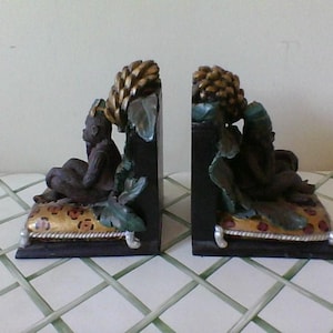 May include: Pair of decorative bookends featuring a seated figure with a gold-colored headdress and a detailed, dark brown finish. The figures rest on a leopard-print cushion with a gold border. The bookends have a black base.