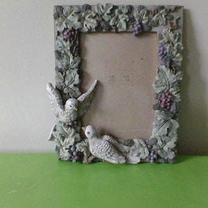 Op de afbeelding: Een wit fotolijstje met een ontwerp van wijnstokken en druiven. Twee witte duiven zitten op het lijstje. Het lijstje staat op een groene ondergrond.