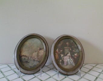 May include: Two oval-shaped framed pictures with ornate gold frames. The picture on the left shows a watercolor painting of a canal scene with buildings. The picture on the right shows a watercolor painting of a group of people in a garden setting.