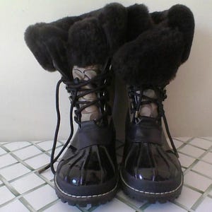 May include: A pair of mid-calf winter boots featuring a black rubberized lower shell, a brown and tan monogram patterned shaft, and thick black faux fur lining at the top. The boots include black laces, metal eyelets, and a lugged sole.