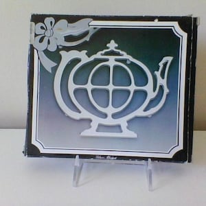 May include: A white ceramic mug with a black handle and the text "But First, Coffee" in a bold, black font. The mug is set against a blurred background, suggesting a cozy setting.