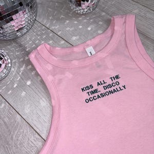 May include: A light pink sleeveless tank top with a crew neckline. The chest area features black embroidered text that reads: KISS ALL THE TIME. DISCO OCCASIONALLY. The garment is made of a solid-colored fabric and includes a small white brand label.